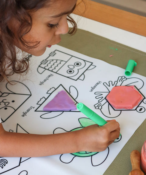 A young child shapes colored dough on a reusable silicone HeyDoodle mat, matching a triangle outline among playful labeled animals.