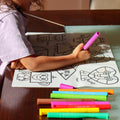 A child colours geometric animals on a reusable HeyDoodle silicone mat, surrounded by bright markers atop a dark wooden table.