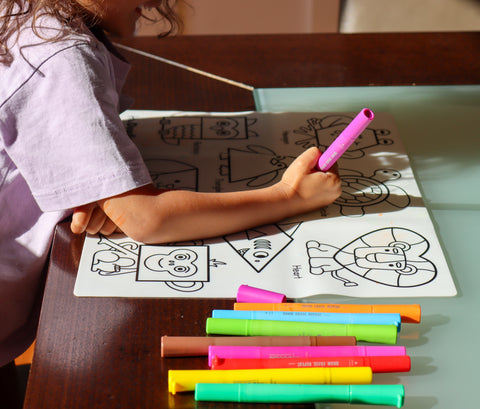 A child colours geometric animals on a reusable HeyDoodle silicone mat, surrounded by bright markers atop a dark wooden table.