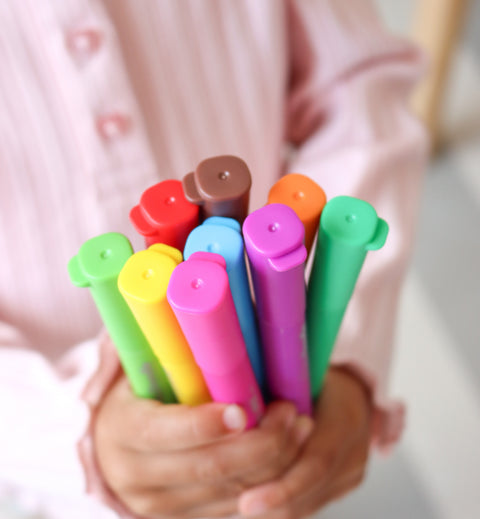 A child in a light pink shirt holds vibrant crayon pens with multicolored caps, perfect for reusable colouring.