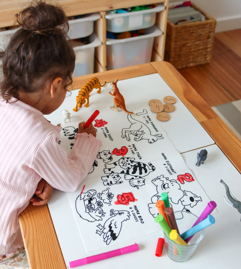 A young child colours animal counting mats at a table with crayon pens, surrounded by markers and animal figurines.