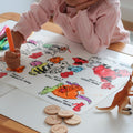 A child in a pink shirt draws with washable crayons on a silicone HeyDoodle counting colouring mat, surrounded by toy animals and wooden discs.