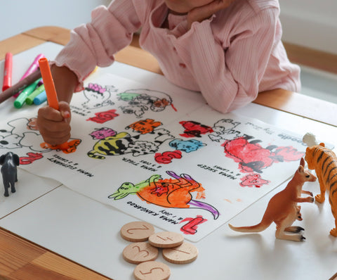 A child in a pink shirt draws with washable crayons on a silicone HeyDoodle counting colouring mat, surrounded by toy animals and wooden discs.