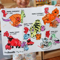 A child holds up a colourful HeyDoodle silicone mat with bright crayon drawings of five animals and their babies, each labeled for counting.