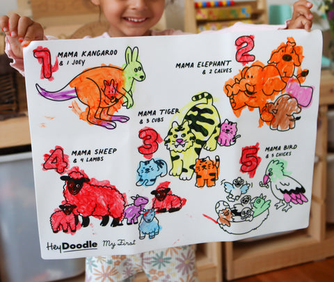 A child holds up a colourful HeyDoodle silicone mat with bright crayon drawings of five animals and their babies, each labeled for counting.