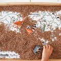 A child’s hand plays with toy dinosaurs on lentils, partly covering colourful animal names and illustrations on a silicone HeyDoodle mat.