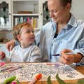 A woman and child colour a silicone HeyDoodle Flower Power mat with markers, smiling amid green leaves and pink florals.