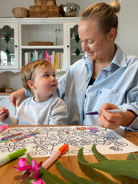 A woman and child colour a silicone HeyDoodle Flower Power mat with markers, smiling amid green leaves and pink florals.