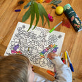 A child colours HeyDoodle’s silicone Flower Power Mini Mat at a wooden table with markers, leaves, a lemon, petals, and more.