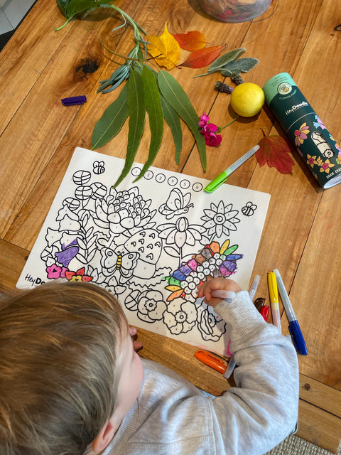 A child colours HeyDoodle’s silicone Flower Power Mini Mat at a wooden table with markers, leaves, a lemon, petals, and more.