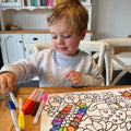 A young child smiles while colouring Australian botanicals on a colourful HeyDoodle silicone mat at a wooden table; books behind.