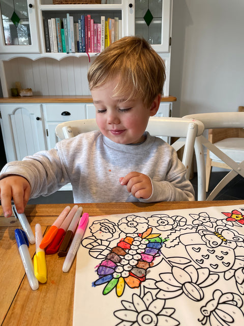 A young child smiles while colouring Australian botanicals on a colourful HeyDoodle silicone mat at a wooden table; books behind.